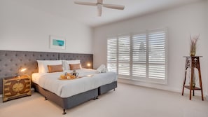 Ein Schlafzimmer mit einem Bett, Nachttischen, einem Deckenventilator, einem Fenster mit Jalousien und einer Zimmerpflanze.