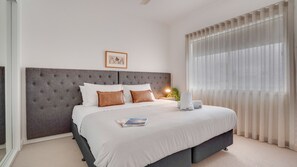 Una habitación de hotel con una cama grande, un cabecero, una mesita de noche con una lámpara y cortinas traslúcidas.