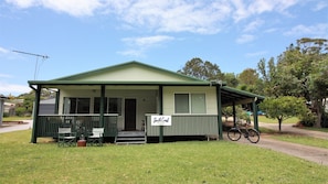 A small, single-story building with a green roof and a porch. There is a sign on the porch that reads 'South Coast Holidays.' - Cherry Street Cottage (Bendalong)