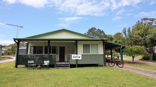A small, single-story building with a green roof and a porch. There is a sign on the porch that reads 'South Coast Holidays.' - Cherry Street Cottage (Bendalong)