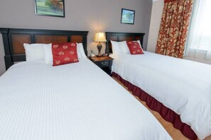 Quarto de hotel com duas camas, cada uma com roupa de cama branca e almofadas vermelhas, uma mesa de cabeceira com abajur e uma janela com cortinas.