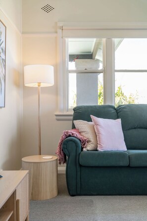 A room with a green sofa, a round side table, a floor lamp, and a window with a view of greenery. - Pearl Cottage – Charming Coastal Retreat (Ulladulla)