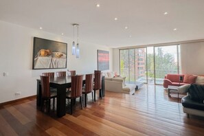 A spacious living area with a dining table and chairs, a sofa, and a large window offering a view of buildings. - Bright 2BR Apartment in Virrey with Private Jacuzzi & AC (Bogotá)