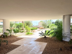 An outdoor covered patio area with a tiled walkway, surrounded by various plants and trees.