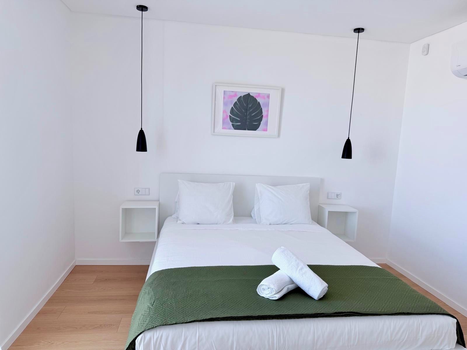 Habitación de hotel minimalista con una cama individual, ropa de cama blanca, una manta verde y una obra de arte enmarcada en la pared.
