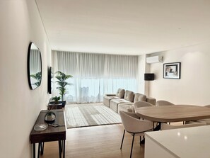 A modern living room with a sofa, a dining table, and a large window with sheer curtains.