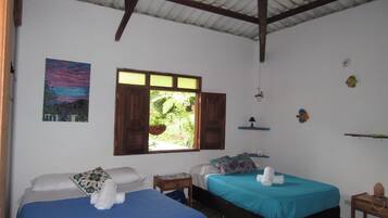 Quarto com duas camas, janela com vista para o verde, um quadro na parede e um pequeno estante com objetos decorativos.