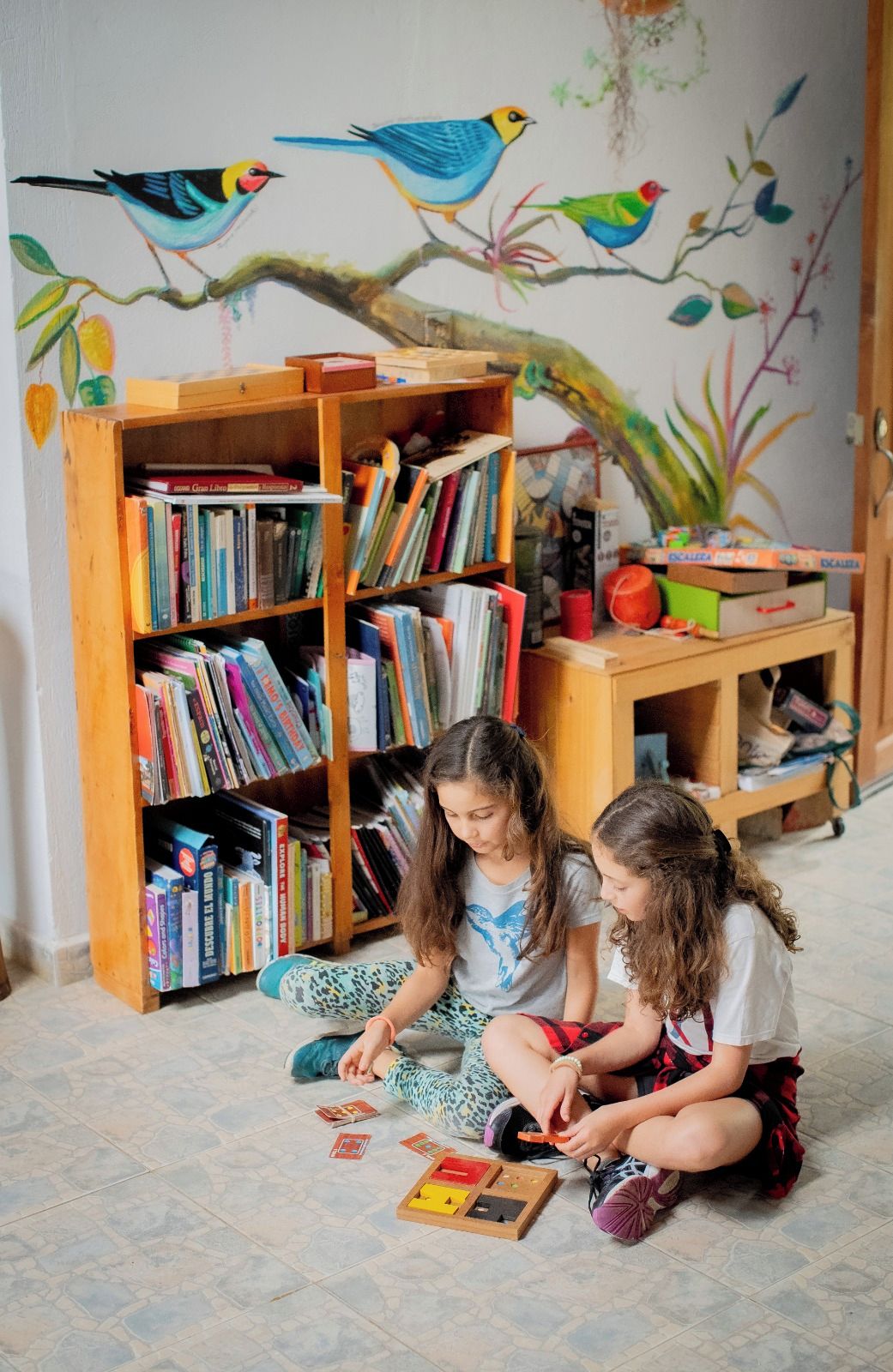 Due bambini che giocano con i giocattoli su un pavimento piastrellato, circondati da scaffali pieni di libri e un murale con uccelli sulla parete.