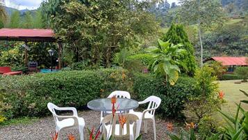 Un jardín con una mesa y sillas, rodeado de diversas plantas y una estructura con techo rojo.