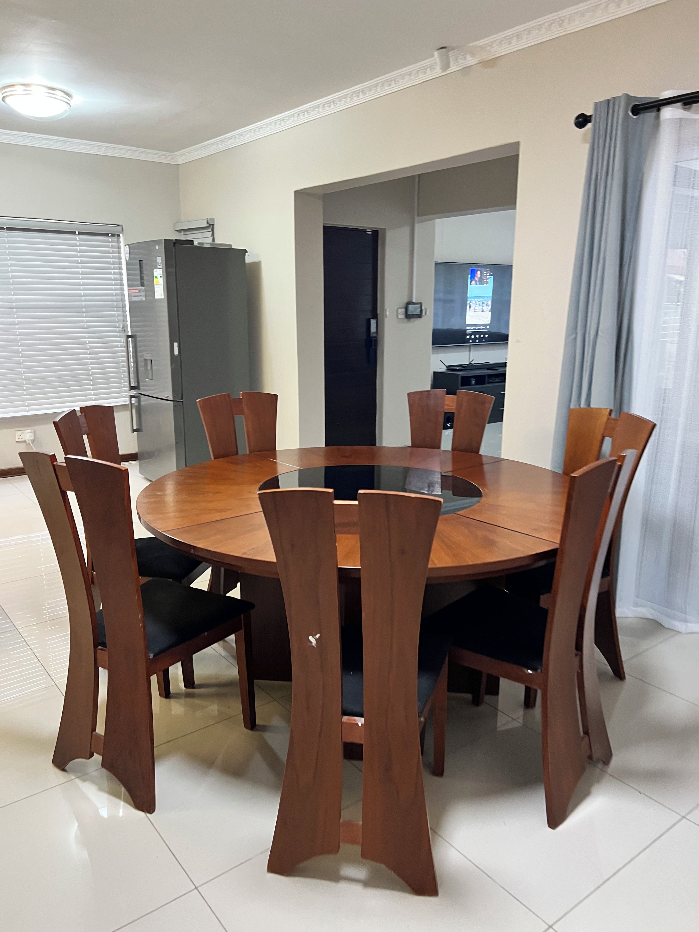 A dining area with a round table and eight chairs, a refrigerator, and a television in the background.
