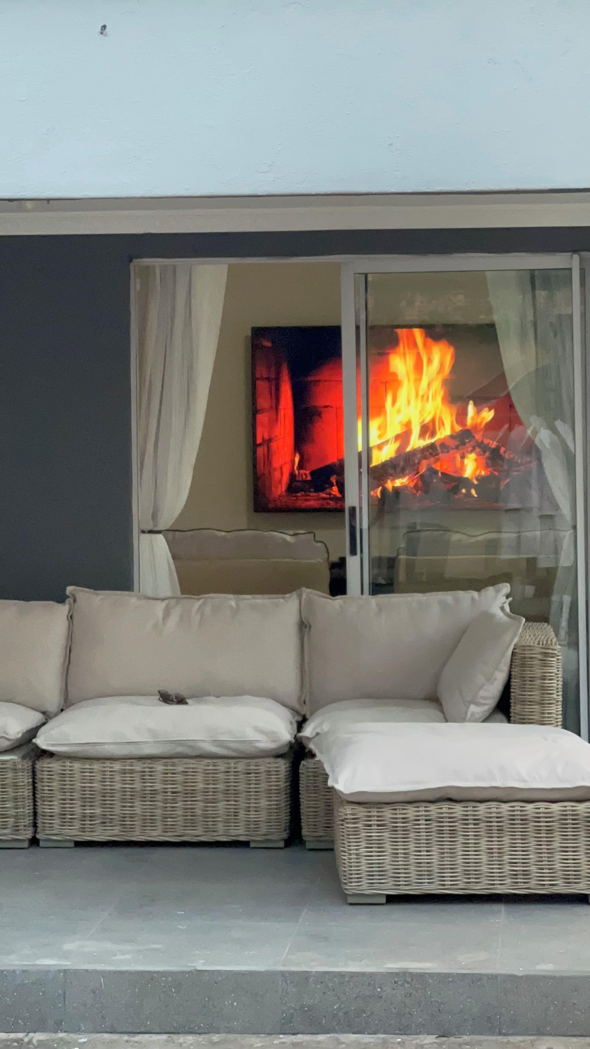 Un canapé en osier avec des coussins, une porte en verre et un feu qui crépite à l’intérieur.