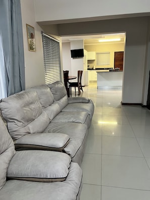 Ein Wohnzimmer mit grauem Sofa, einem Esstisch mit Stühlen und einer Küchenzeile im Hintergrund.