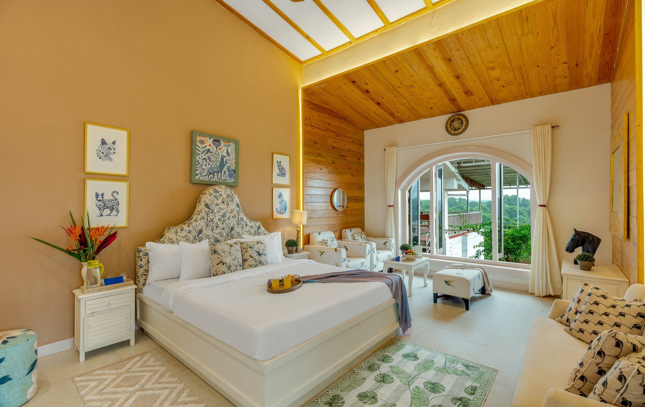 Un dormitorio con una cama grande, un rincón de descanso con sofá y sillones, un ventanal con vistas a zonas verdes y un techo de madera.