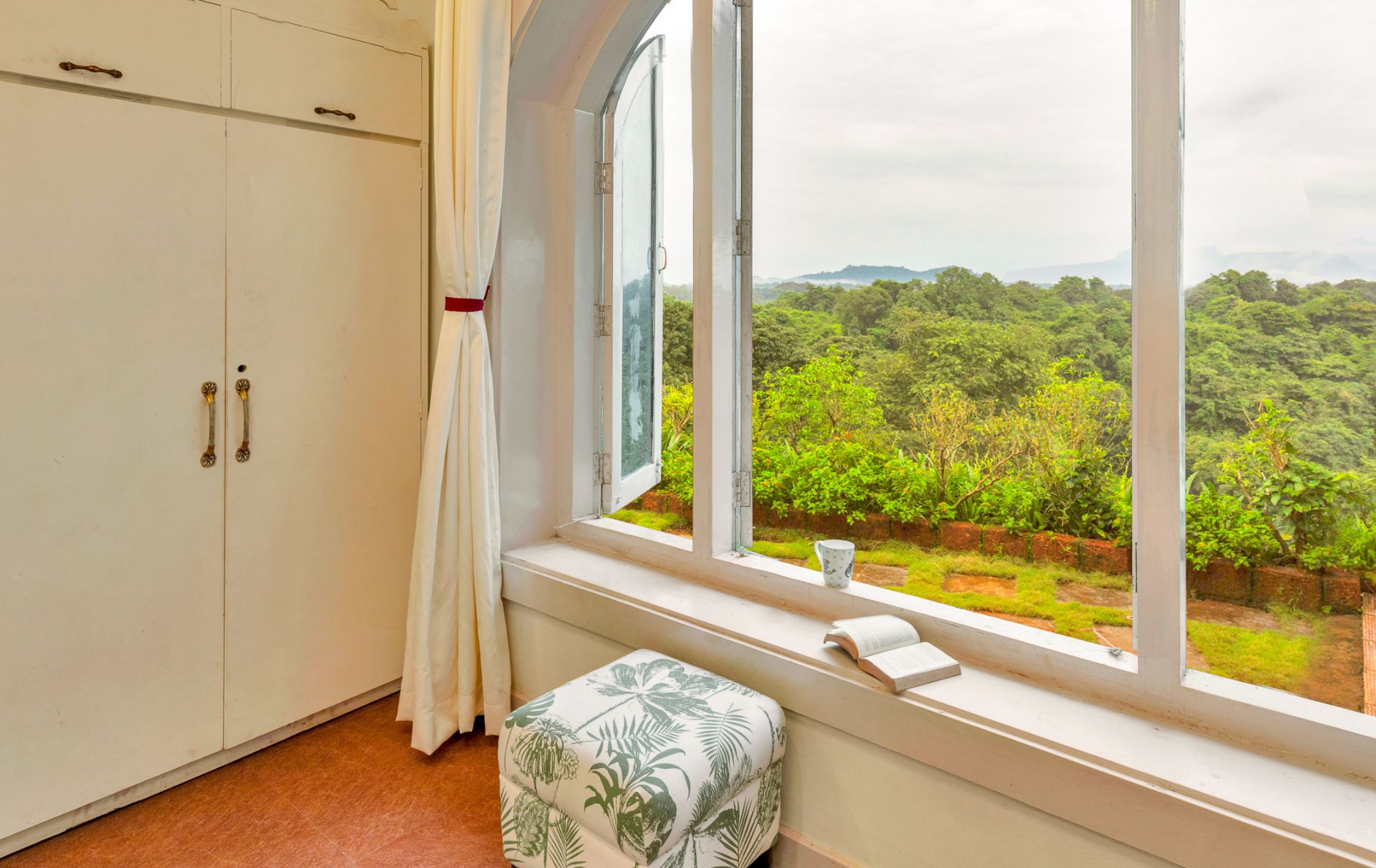 A room with a large window, a white wardrobe, a bench with a tropical print, and a book on the windowsill.