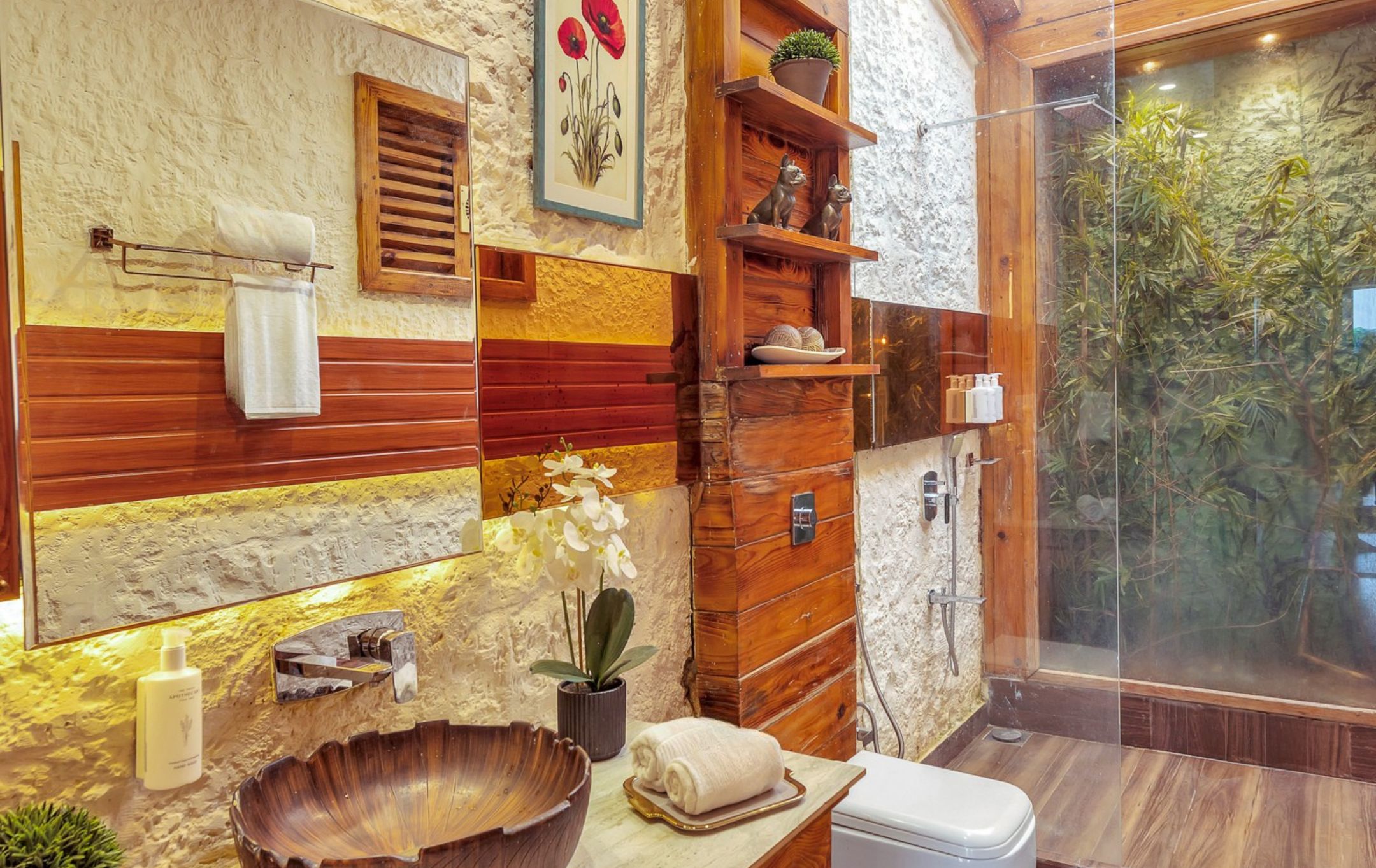 A bathroom with a stone wall, wooden paneling, a large mirror, a sink with a shell basin, and a glass shower enclosure with a view of greenery.