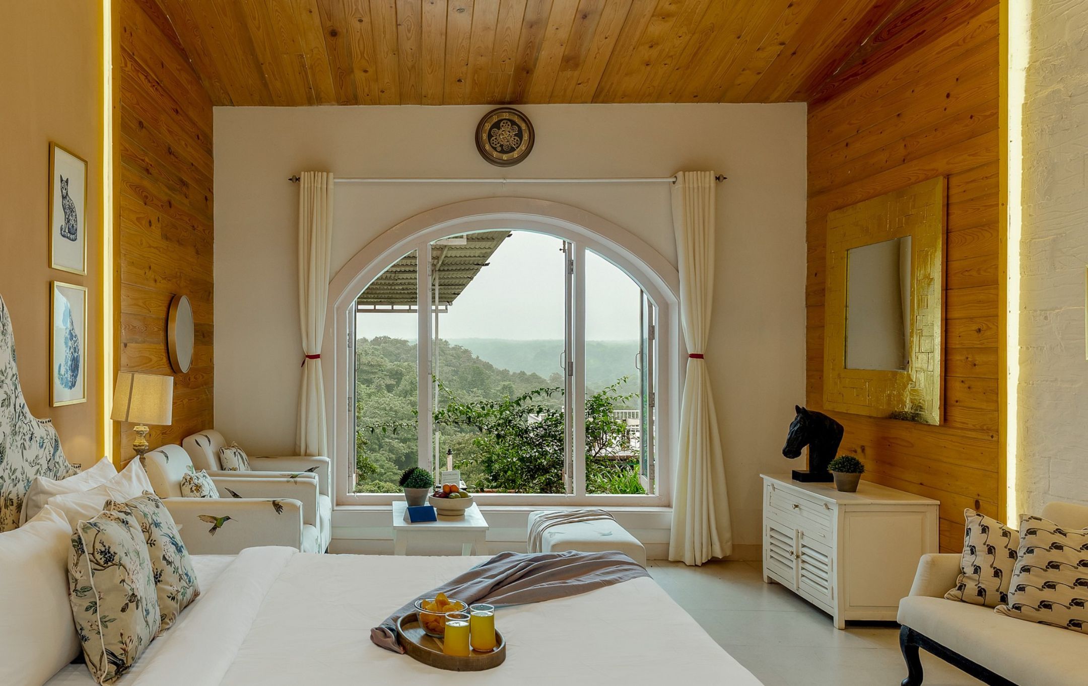 A bedroom with a large window, a bed, a sofa, and a side table with a tray of fruit and juice.