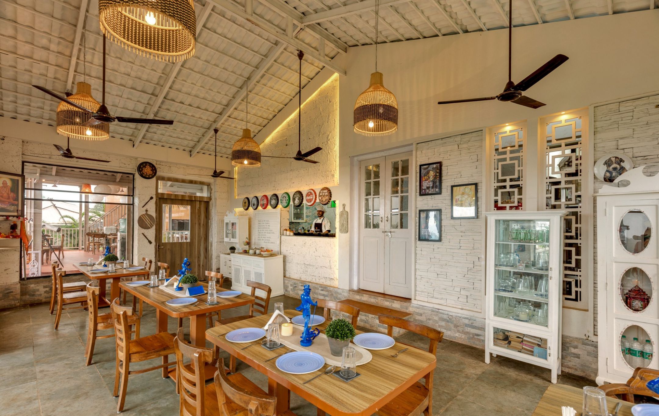A restaurant with wooden tables and chairs, hanging wicker chandeliers, and a white brick wall with framed pictures.