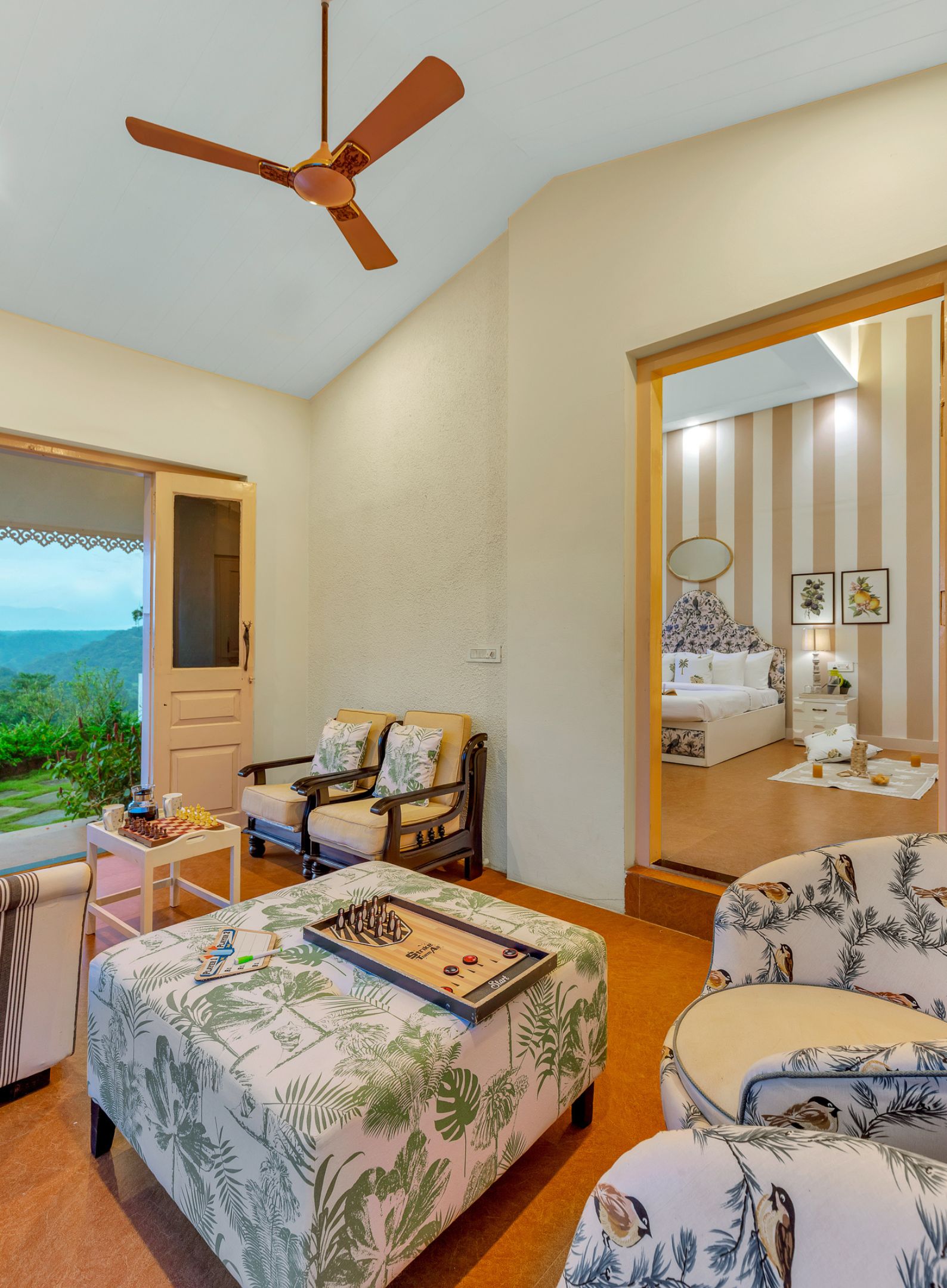 A bedroom with a bed, a chair, a table with a board game, a ceiling fan, and a view of the outdoors.