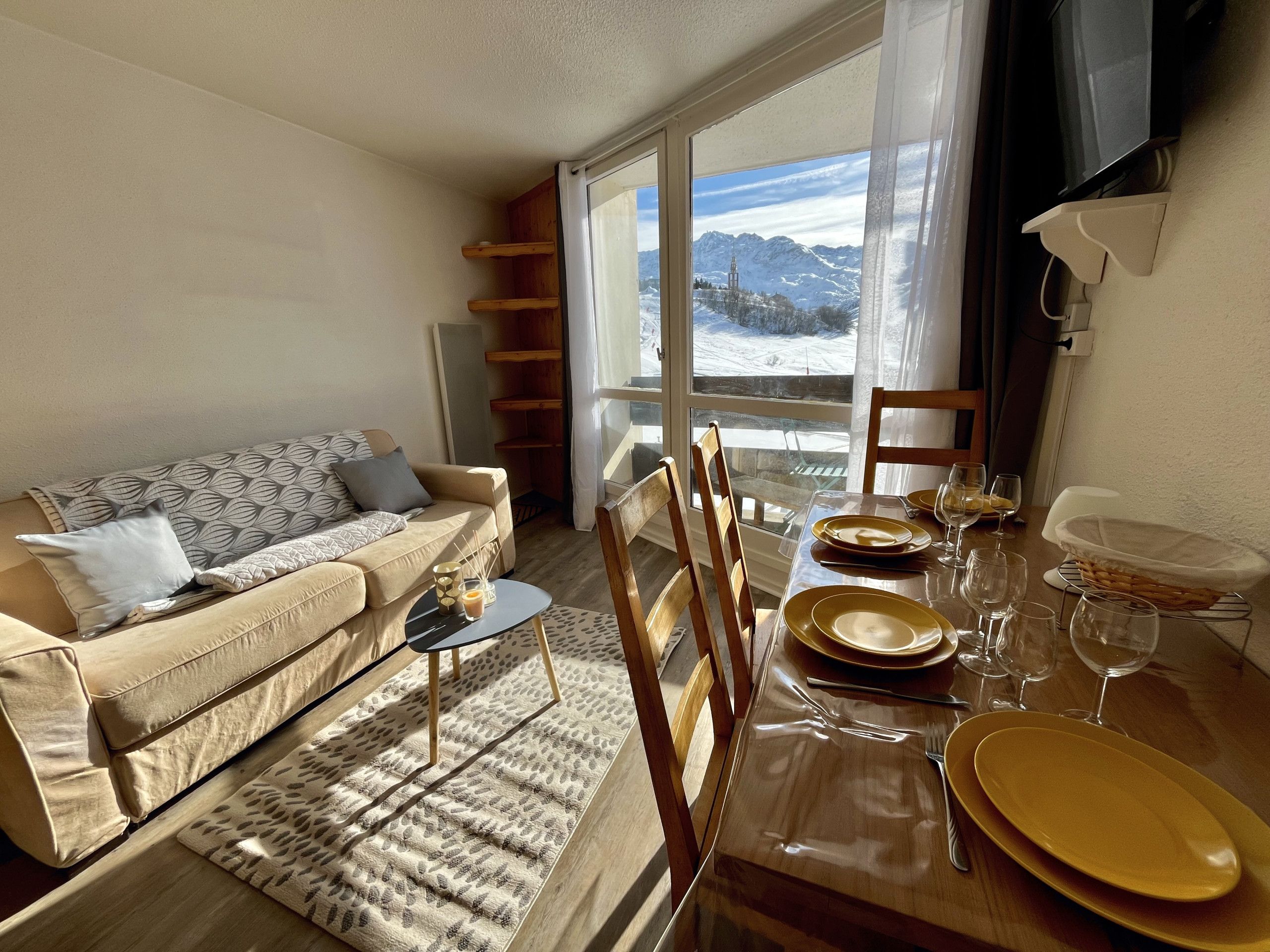 A living room with a sofa, a dining table set with plates and glasses, and a view of snow-covered mountains through large windows.
