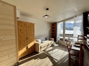 A cozy living room with a wooden wardrobe, a sofa, a dining table with chairs, and a view of a snowy landscape through large windows. - Estrella - Les 3 Vallées - Les Ménuires (Les Menuires)