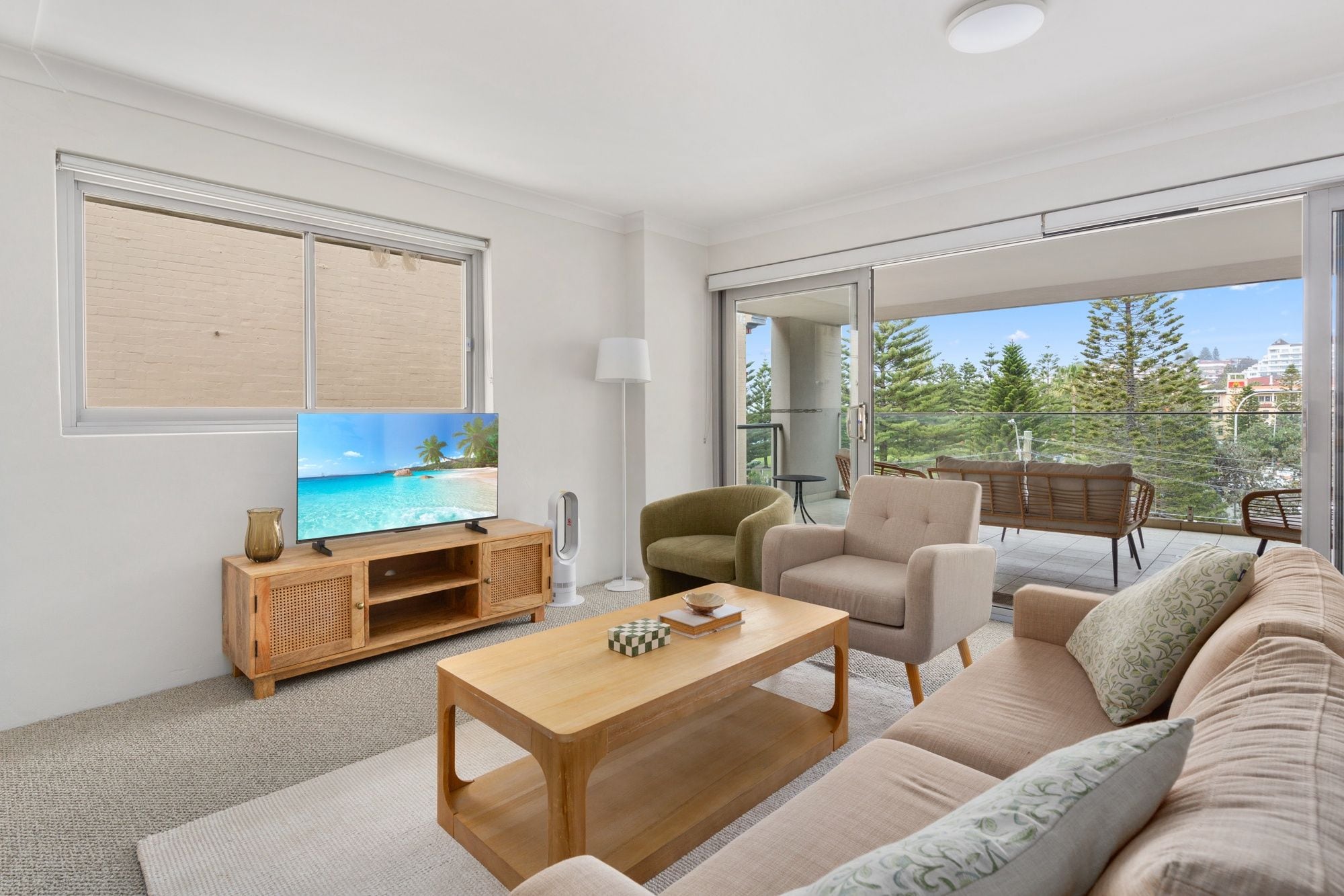 A modern living room with a flat-screen TV, wooden entertainment unit, sofa, and a balcony with outdoor furniture.