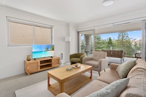 Uma sala de estar moderna com televisão de tela plana, móvel para TV em madeira, sofá e uma varanda com mobiliário de exterior.
