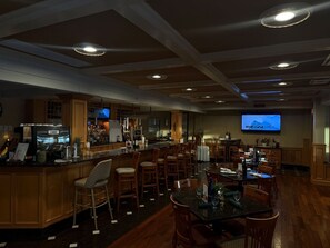 Área de bar com mobiliário de madeira, uma televisão na parede e balcão com diversos objetos.