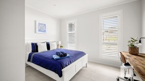 Quarto com cama, escrivaninha e duas janelas com persianas.