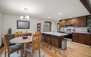 A modern kitchen with wooden cabinets, a stainless steel refrigerator, and a dining area with a round table and chairs. - Lark & Leisure (Bend)