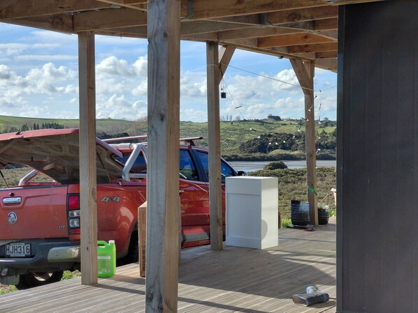 A covered outdoor area with a red truck parked under a wooden structure, a white utility box, and a view of a lake and hills in the background.