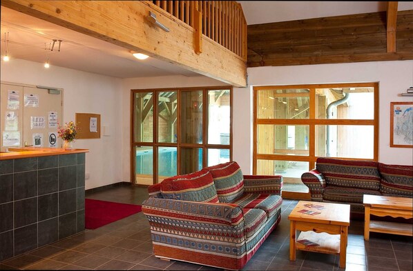Un hall espacioso con techo alto, vigas de madera, un mostrador de recepción, sofás estampados y ventanas grandes.
