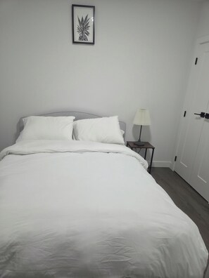 A neatly made bed with white bedding, a framed pineapple artwork on the wall, and a bedside table with a lamp. - Cozy 2-bedroom basement apartment (Calgary)