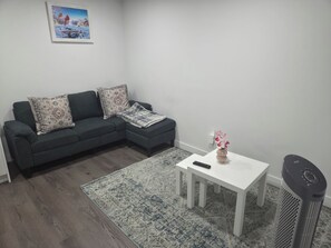 A living room with a dark grey sofa, patterned cushions, a white coffee table with a vase of flowers, a remote control, and a standing heater. - Cozy 2-bedroom basement apartment (Calgary)