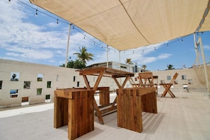 Un espacio de estar al aire libre con mesas y sillas de madera bajo un toldo grande, rodeado por un muro de concreto con ventanas.