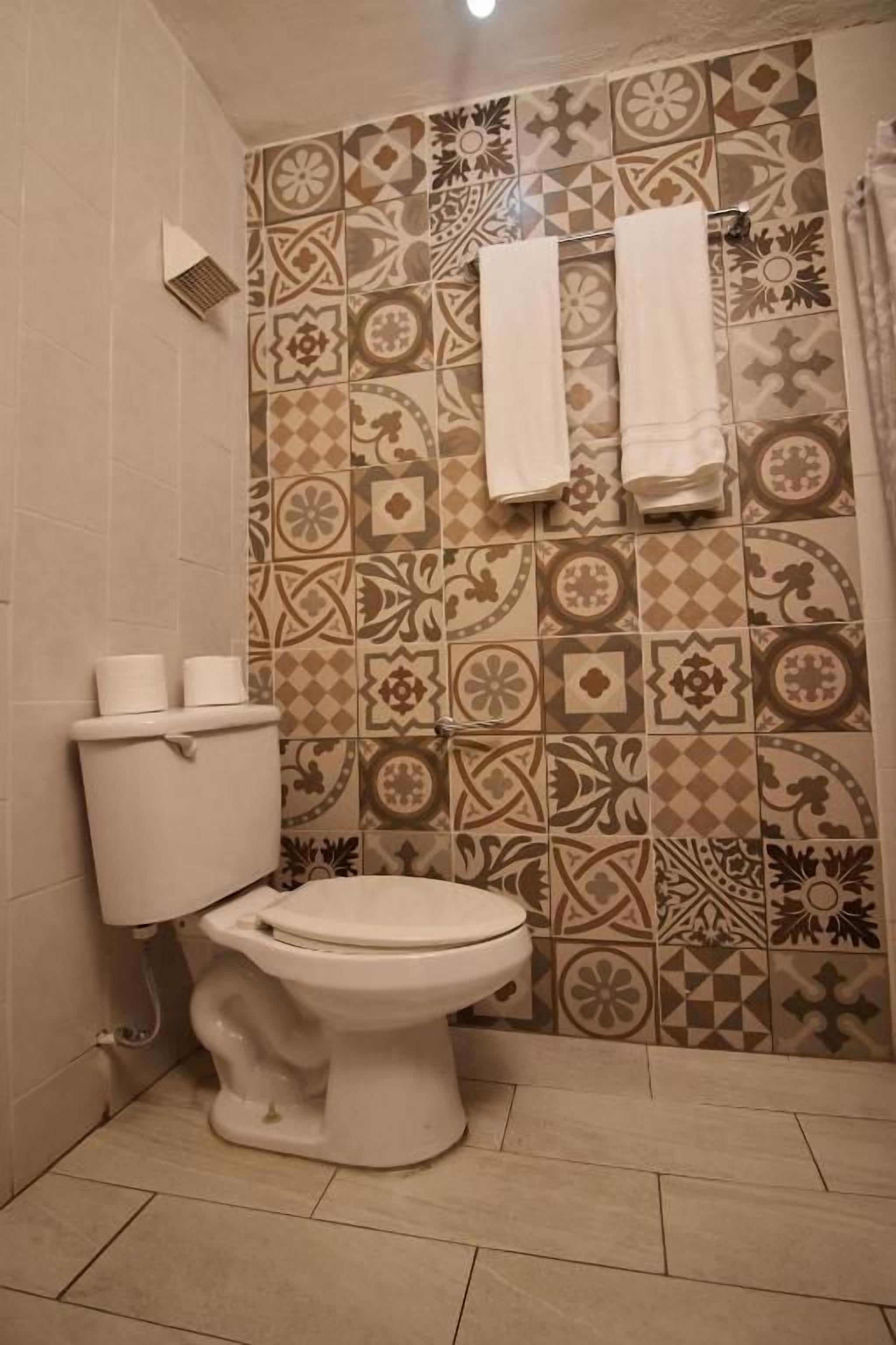 Un baño con inodoro, baldosas estampadas y dos toallas blancas colgadas en la pared.