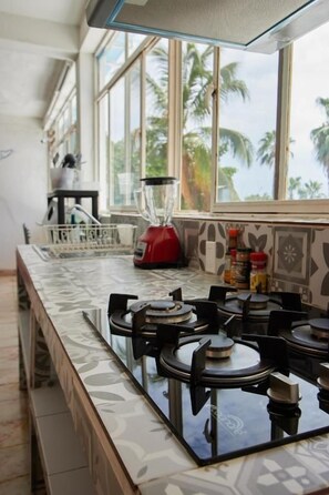 Una cocina con estufa a gas, licuadora y ventana con vista a palmeras.