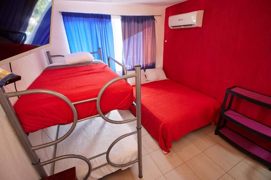 Een kamer met twee stapelbedden van metaal, een rode wand, een raam met blauwe gordijnen en een witte airconditioning.