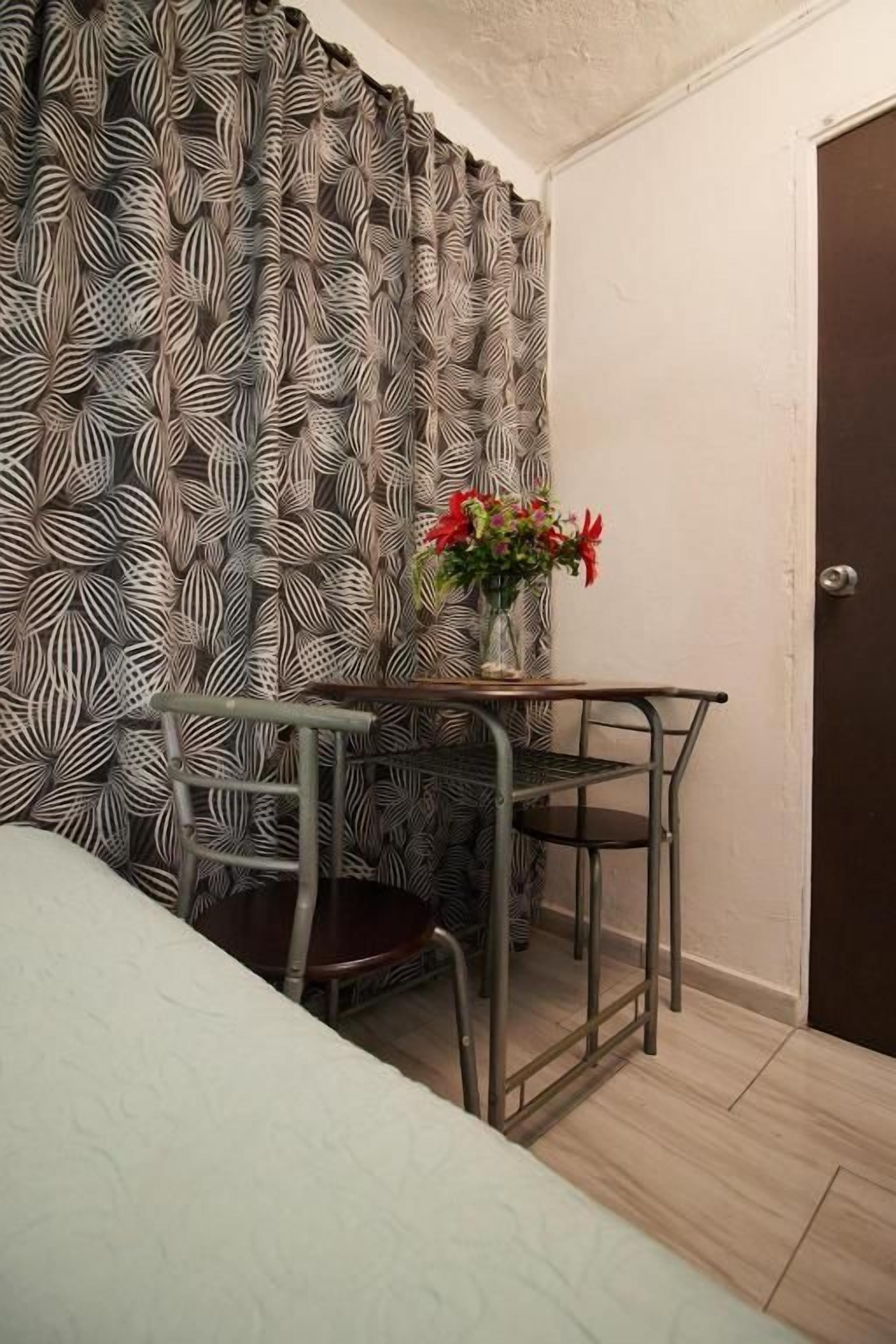 Una habitación pequeña con una cama, un escritorio con una silla, un florero con flores y una cortina estampada.