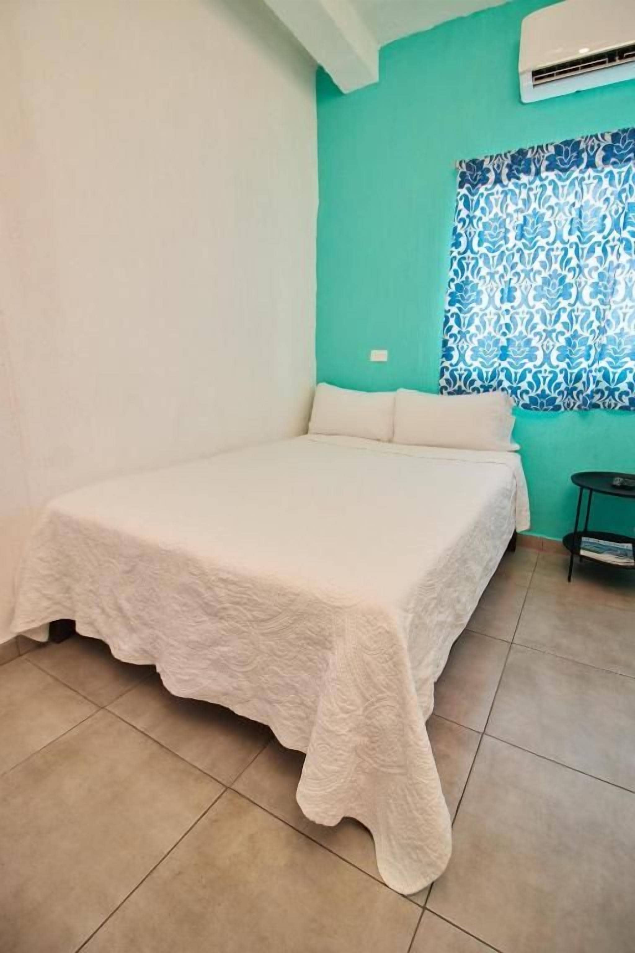 Una habitación con cama, una ventana con cortinas estampadas y una mesita con una revista.