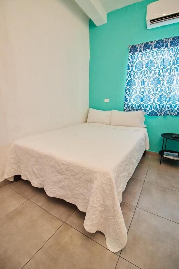 Una habitación con cama, una ventana con cortinas estampadas y una mesita con una revista.