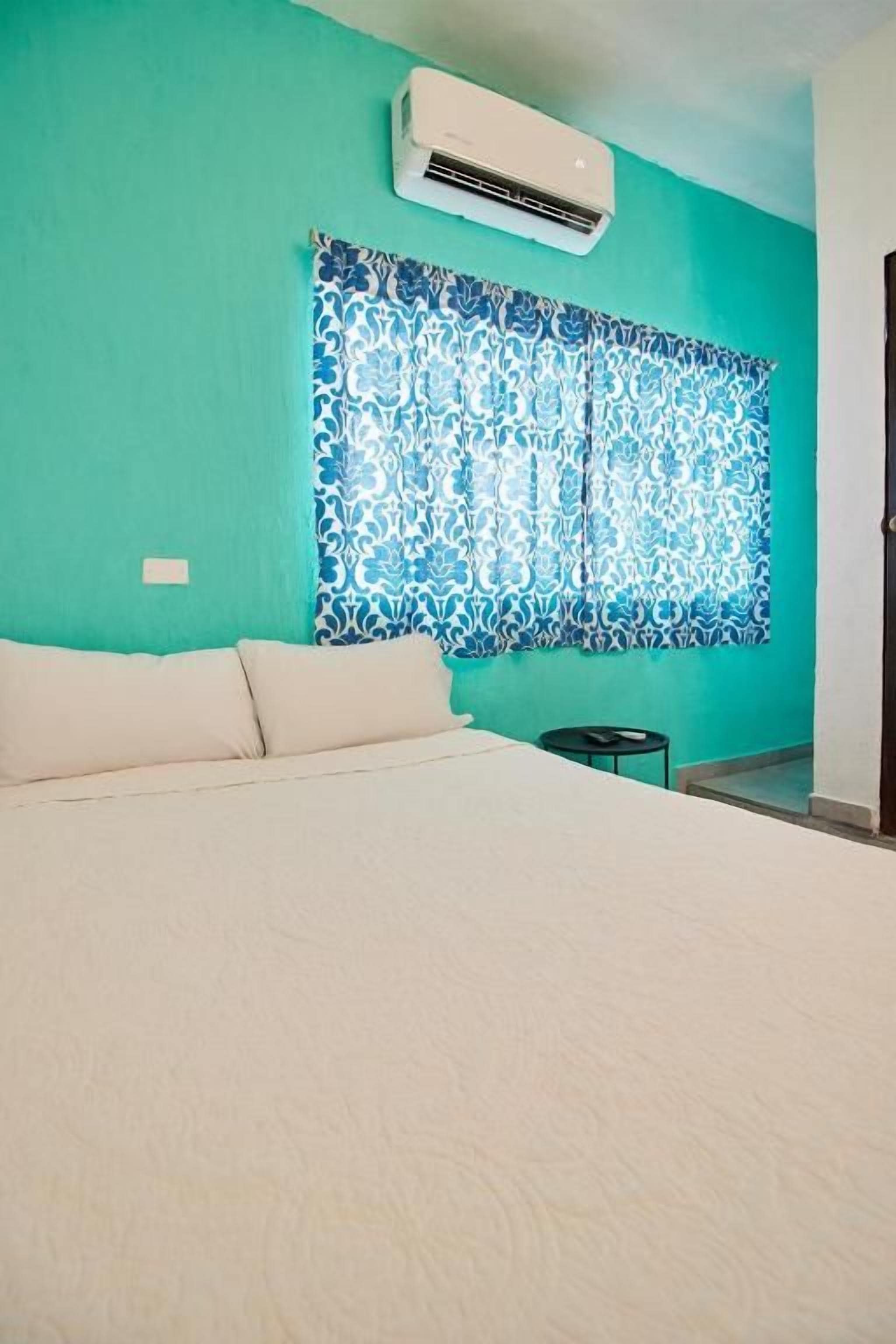 A bedroom with a bed, a window with patterned curtains, an air conditioning unit, and a small bedside table.