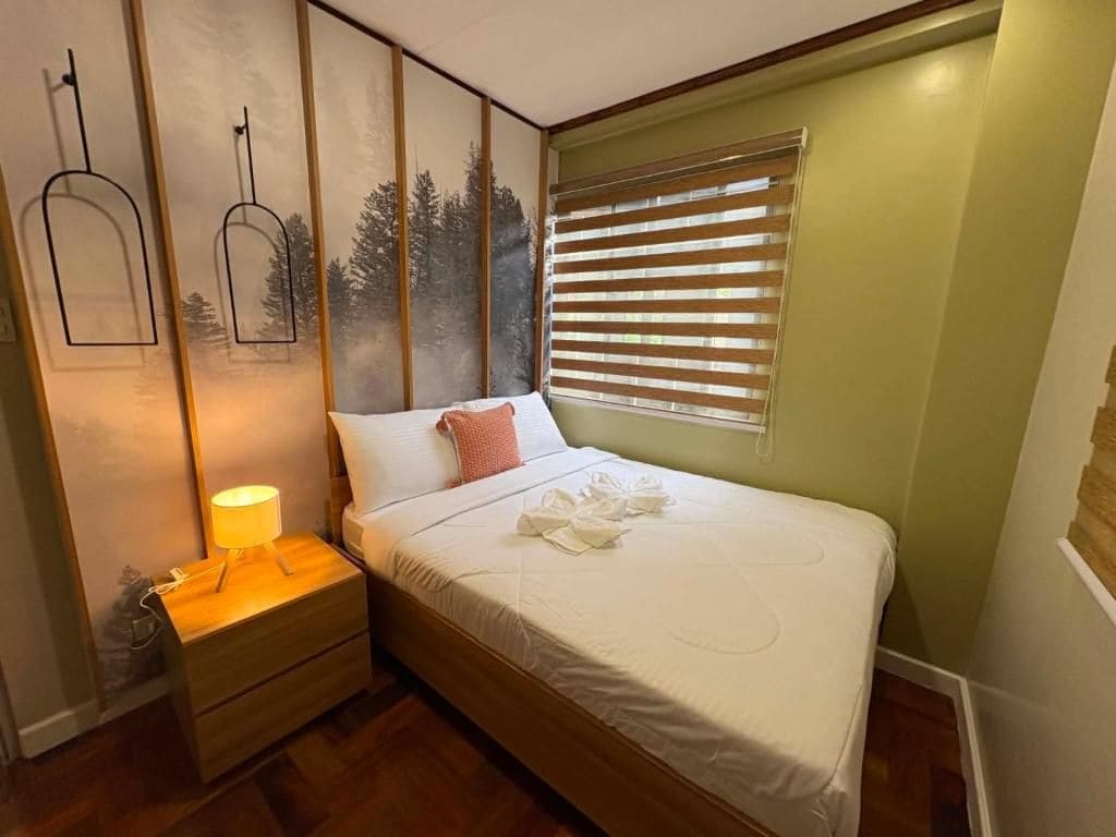 Una cama bien hecha con edredón blanco y una almohada, una mesita de noche con lámpara y una ventana con persianas.