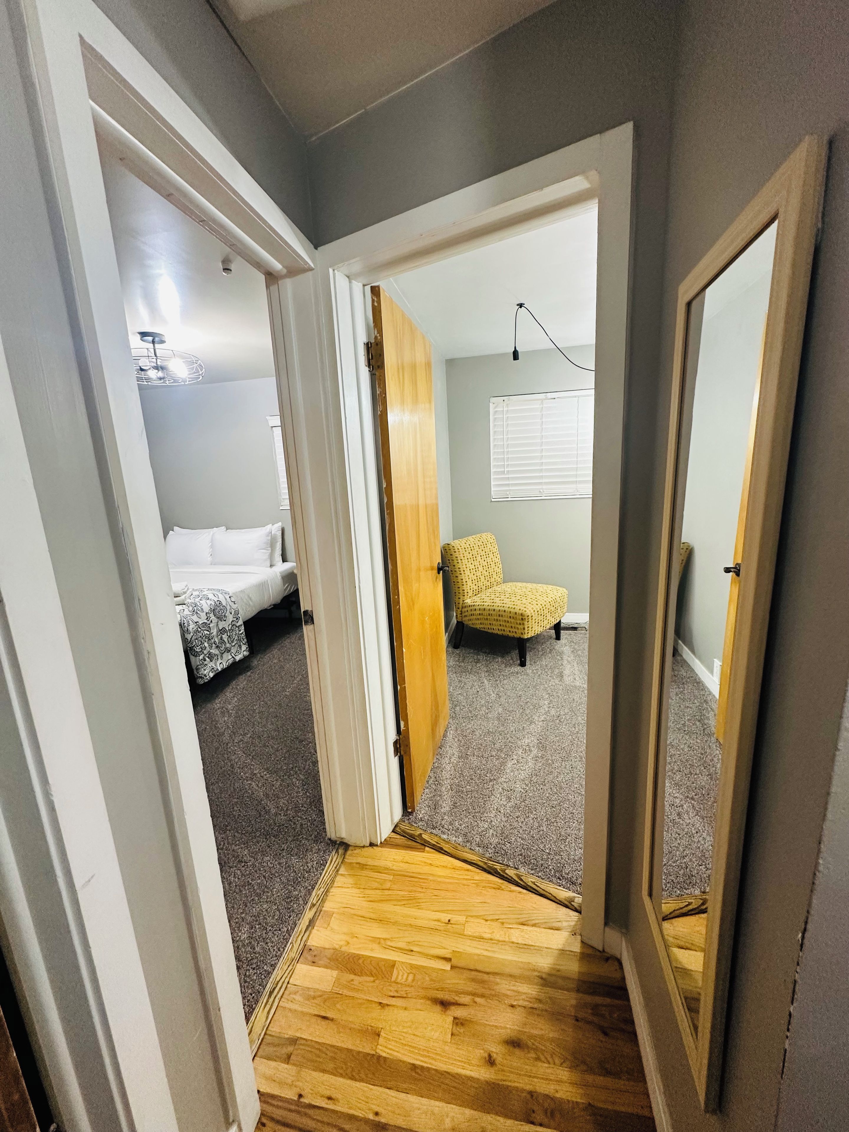A narrow hallway leading to a bedroom with a bed, a yellow chair, and a wooden floor.