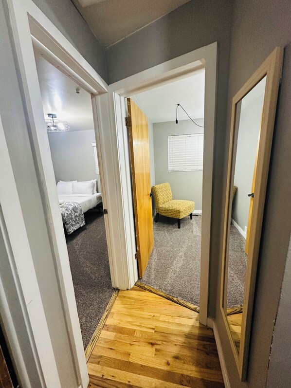 Un couloir étroit mène à une chambre où se trouvent un lit, une chaise jaune et un plancher en bois.
