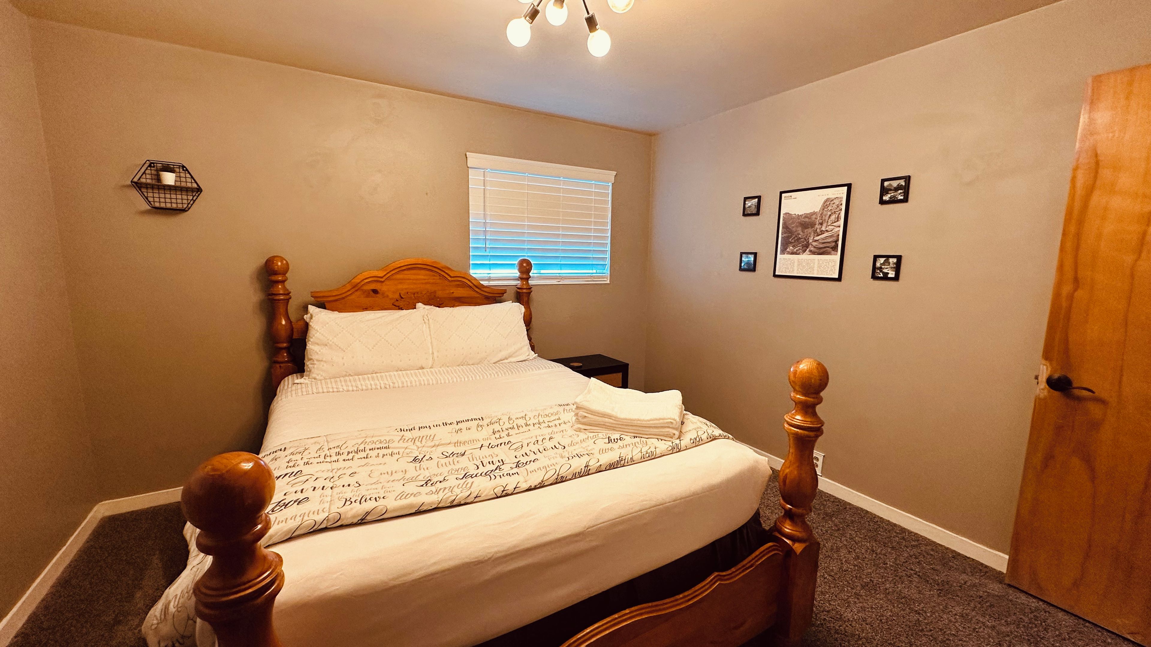A bedroom with a four-poster bed, a window with blinds, a small bedside table, and a door.