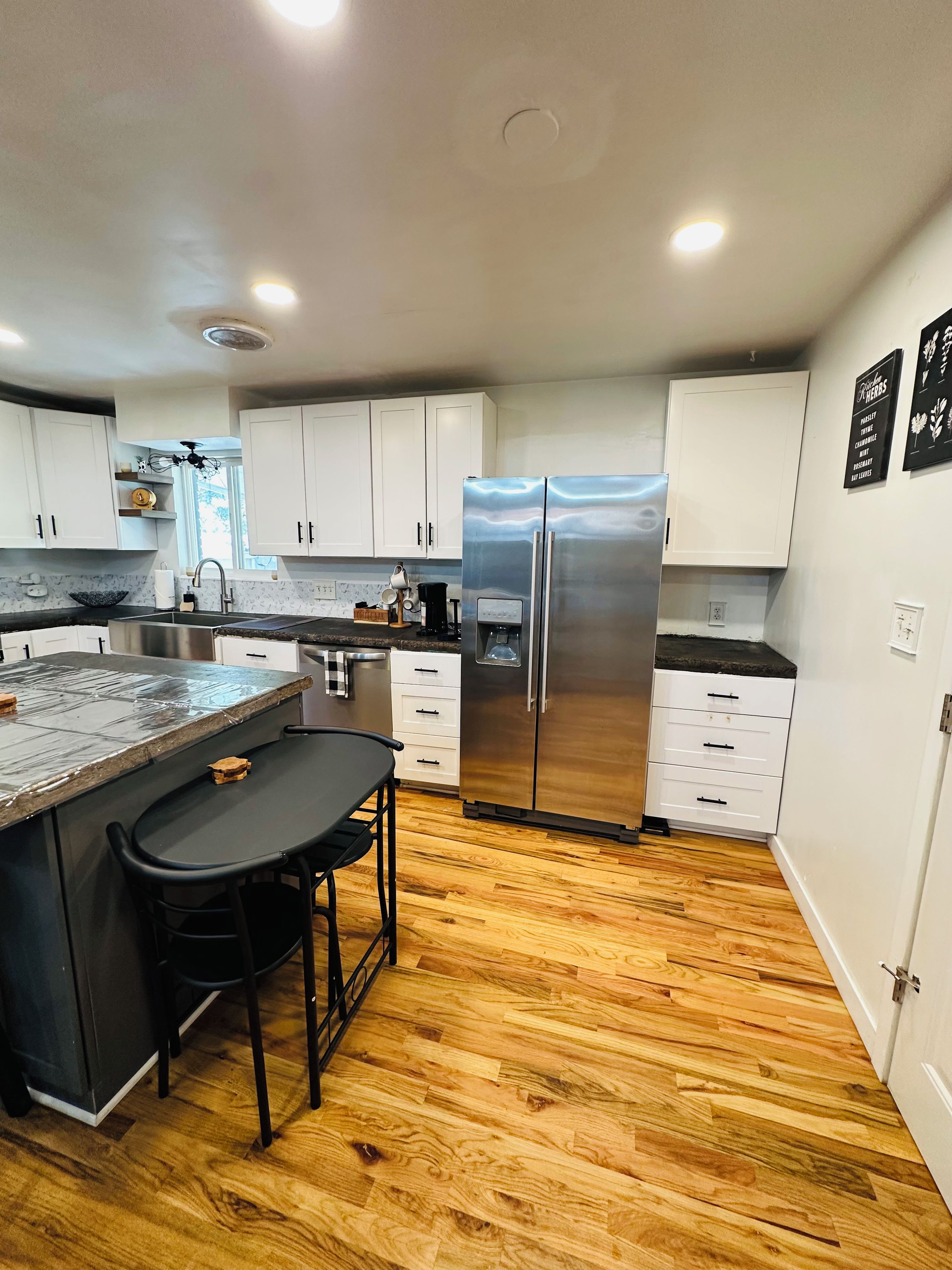 A modern kitchen with white cabinets, a stainless steel refrigerator, a bar area with stools, and wooden flooring.