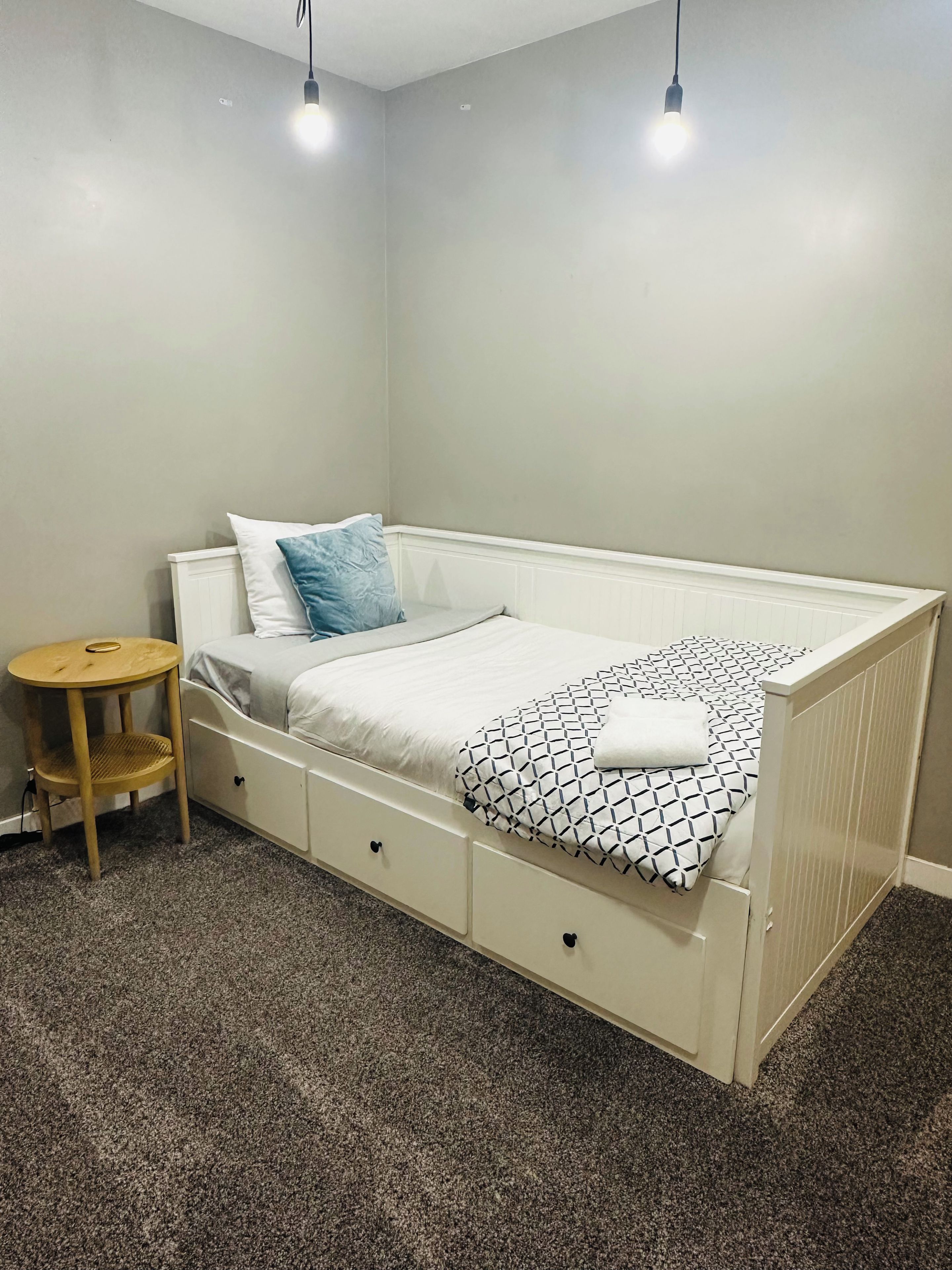 Une chambre avec un lit simple, une tête de lit blanche, une petite table de chevet en bois et une table ronde avec un siège en osier.