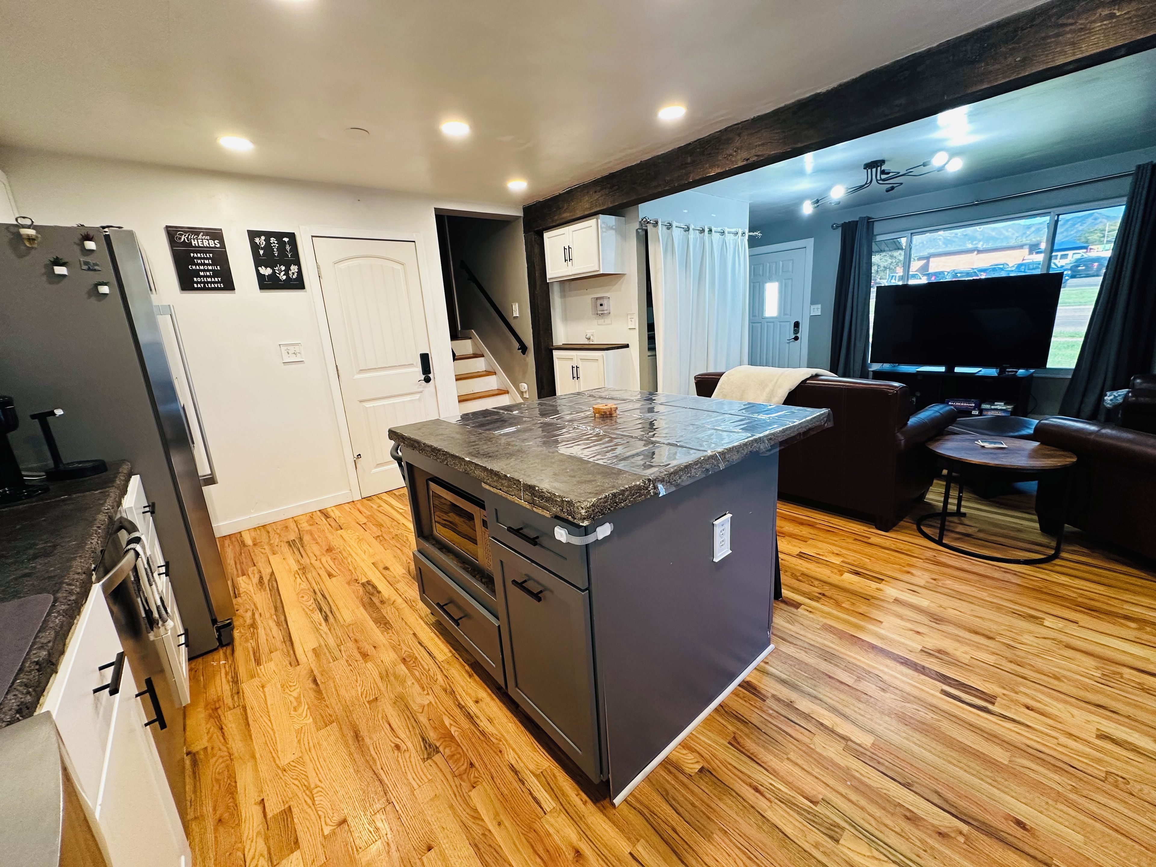 A modern kitchen with a central island, wooden flooring, and a view of the living area with a TV and seating.