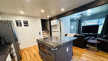 A modern kitchen with a central island, wooden flooring, and a view of the living area with a TV and seating.