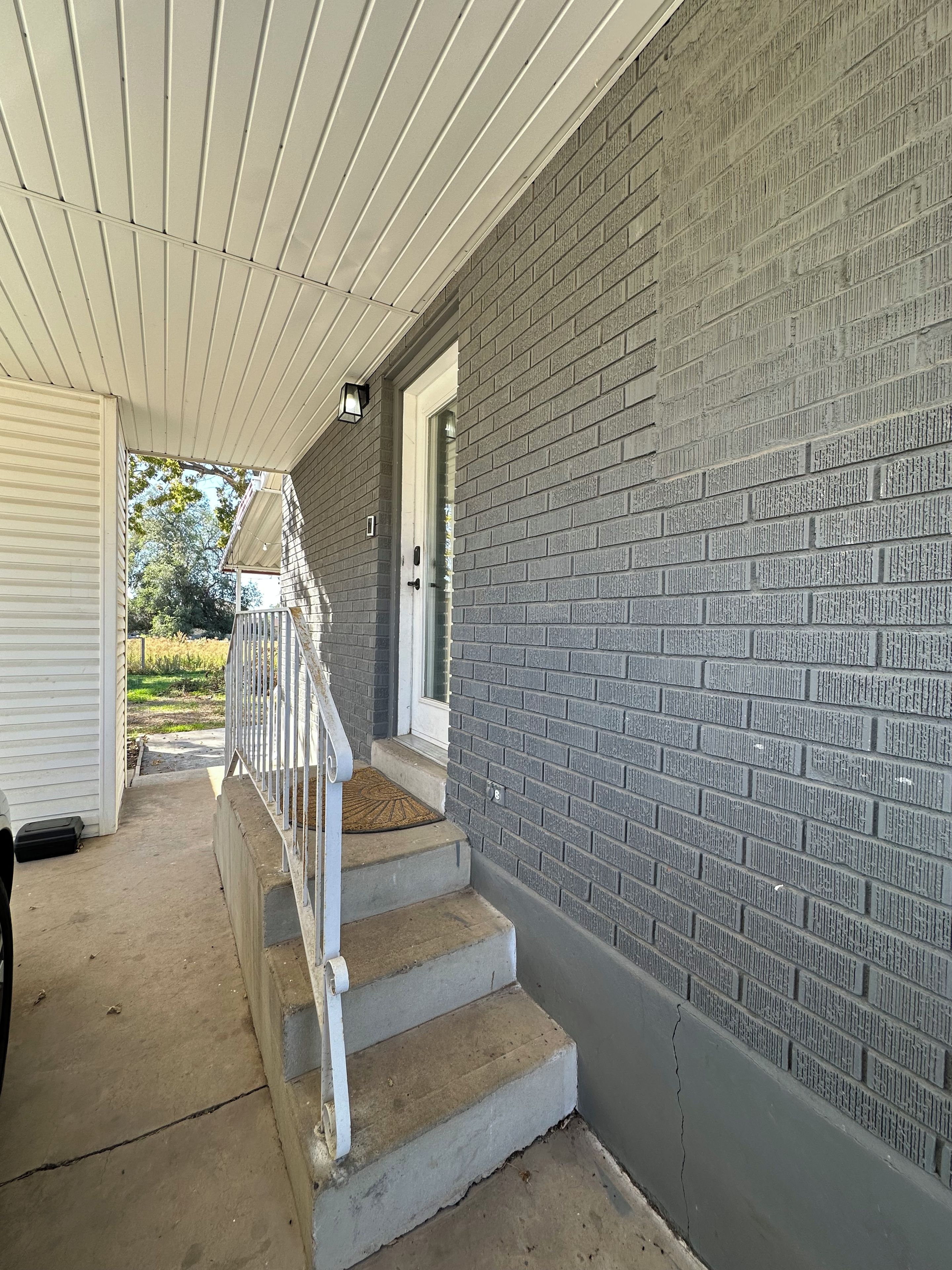 Une véranda couverte avec un mur en briques, une balustrade blanche et une porte donnant sur l’intérieur.